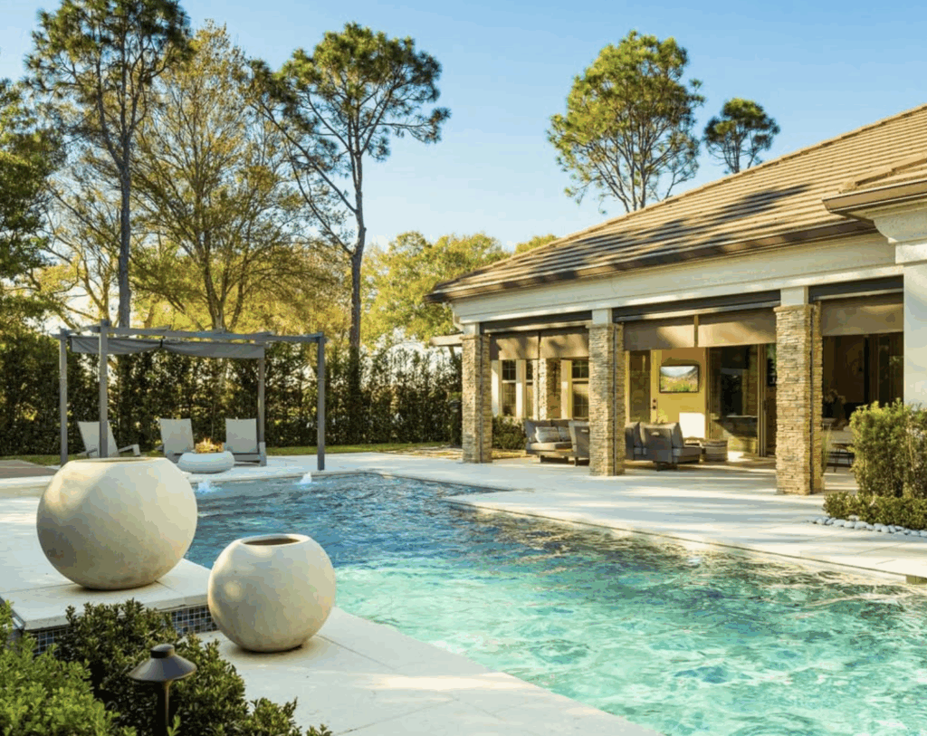 Modern backyard with a rectangular swimming pool, large round planters, lounge chairs under a pergola, and a patio attached to a house with stone columns.